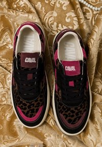 Paire de baskets noires, marron imprimé léopard et roses avec l’étiquette "CAVAL" sur la languette, posées sur un tissu à motif doré avec un collier de perles à proximité.