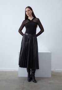 Juodas ilgomis rankovėmis megztinis su kryžiaus dizainu, derinamas su raukšlėtu juodos odos sijonu ir juodais aukštakulniais iki kelių, pastatytas neutralaus fono kontekste.