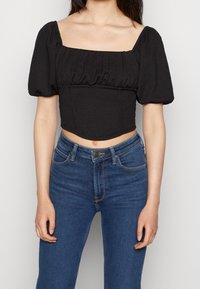 Svart kort puffärmad topp med fyrkantig halsringning, texturerad tyg och rynkad detalj runt bysten, parat med blå högmidjade jeans.