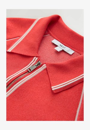 Červená polokošeľa so zipsovým zapínaním, biele pruhy pozdĺž goliera a zipsu, textúrovaný materiál a štítok s nápisom "NEXT".