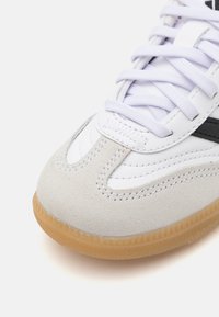Scarpa atletica bianca con punta in suede grigia, strisce laterali nere e suola in gomma naturale con dettagli testurizzati. I lacci sono bianchi.