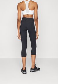 Schwarze Capri-Leggings aus dehnbarem Stoff, mit hohem Bund und kleinem Logo auf der Rückseite, kombiniert mit einem weißen Sport-BH.