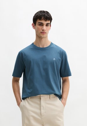 Młody mężczyzna o krótkich ciemnych włosach, ubrany w turkusowo-niebieski T-shirt z krótkim rękawem i beżowe spodnie, stojący z rękami w kieszeniach.