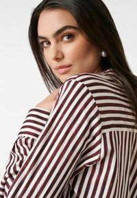 Woman with long dark hair wearing a maroon and white striped shirt, looking over her shoulder with a neutral expression.