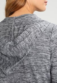 Melírovaná šedá mikina s kapucí, vyrobená z měkké, texturované látky, s výstřihem do kulata a uvolněným střihem. Kapuce má jemné detaily prošití.