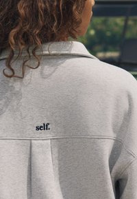 Person mit lockigem Haar trägt ein hellgraues Hemd mit Kragen und dem Wort "self.", das auf den oberen Rücken gestickt ist.