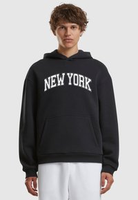 Crna dukserica s kapuljačom s bijelim natpisom "NEW YORK" preko prsnog dijela. Ima prednji džep te rebraste manšete i rub. Mekana tekstura tkanine.