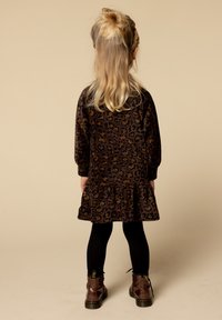 Kind met lang blond haar, gekleed in een bruine jurk met luipaardprint, zwarte leggings en bruine laarzen, staand met de rug naar de camera op een beige achtergrond.