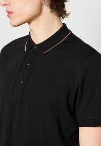 Homme portant un polo noir à trois boutons, avec un discret liseré rose au col et un logo brodé "BOSS" sur la poitrine.