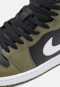 Olive green and black leather sneaker with white Nike logo, textured laces, and a white rubber sole. Features smooth and stitched accents.