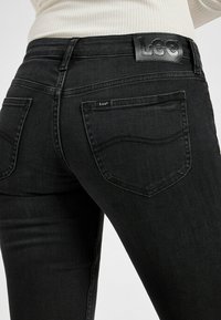 Jean en denim noir avec une coupe ajustée, présentant une patch en cuir avec logo, deux poches arrière et des détails de couture subtils.