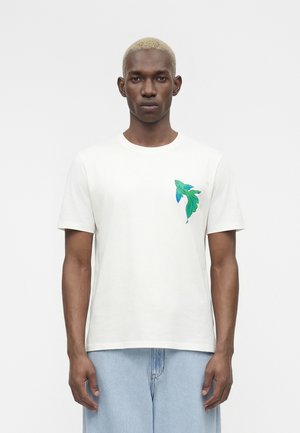 Άνδρας με κοντά ξανθά μαλλιά που φοράει λευκό μπλουζάκι με πολύχρωμο γραφικό από πράσινο και μπλε ψάρι στο αριστερό μέρος του στήθους και ανοιχτό μπλε τζιν.