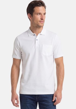 Mann trägt ein weißes, kurzärmeliges Poloshirt mit einer Brusttasche und dunkelblaue Jeans, blickt leicht nach rechts.