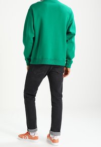 Person som bär en grön sweatshirt, svarta uppvikta jeans, vita strumpor och orangea och vita sneakers, står vänd bort från kameran på vit bakgrund.
