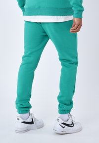 Pantalones de chándal turquesas con puños elásticos, costuras laterales y un ajuste relajado, combinados con zapatillas blancas que tienen un logo de swoosh negro.