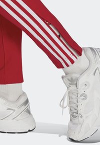 Röda idrottsbyxor med vita sidostråk som har dragkedjor, kombinerat med vita sneakers med silverdetaljer och texturerad nätbeläggning.