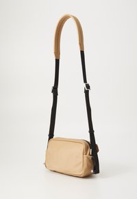 Tan crossbody bag with a smooth texture, zip closure, two side pockets, and an adjustable black strap with a logo pattern.