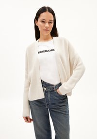 Beige gebreide cardigan met een open voorkant, lange mouwen en een geribbelde textuur, gedragen over een wit shirt met zwarte tekst en blauwe high-waisted jeans.