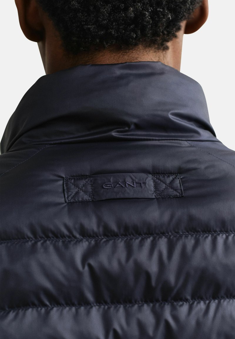 Rückansicht einer Person, die eine dunkelmarineblaue gesteppte Jacke mit hochgestelltem Kragen und einem eingenähten GANT-Label am oberen Rücken trägt.