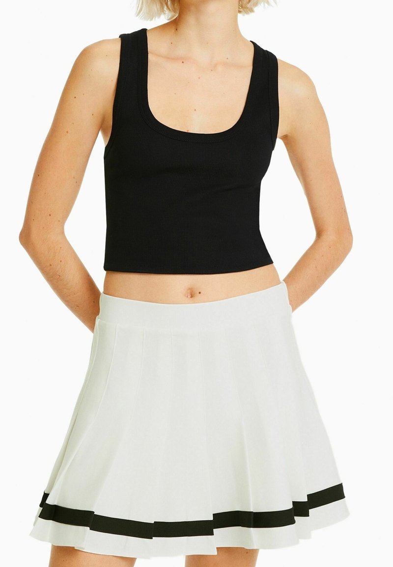 Femme portant un crop top noir sans manches et une jupe plissée blanche avec une bande noire près de l'ourlet, debout les mains derrière le dos.