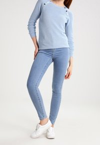 Ljust blå ribbad tröja med knapppdekorer, i kombination med tighta ljusblå jeans och vita sneakers. Slät textur och smal design.