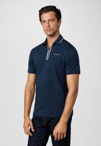 Mornarica polo majica s kratkim rukavima, okovratnikom na zatvarač, bijelim obrubom i logotipom. Glatka tekstura, strukirana kroja i bočni prorezi za udobnost.
