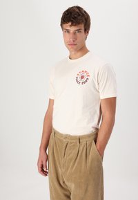 Camiseta corta de manga con estampado floral rojo y texto, combinada con pantalones de pana beige con pliegues frontales y bolsillos.