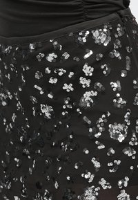 Mustast helvestega seelik läbipaistva pealiskihiga, millel on laiali hõbedased ja mustad ümmargused helbed, rõhutades tekstuurset ja peegeldavat pinda.