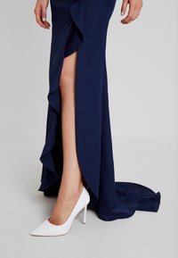 Jambe inférieure d'une femme en robe bleu marine avec une fente haute jusqu'à la cuisse, portant une chaussure blanche à talon aiguilles, debout sur un sol gris clair.