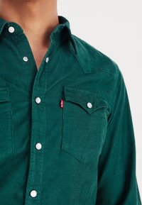Camicia verde in velluto a coste con chiusura frontale a bottoni, colletto appuntito, due tasche sul petto con patte e bottoni a pressione argentati.