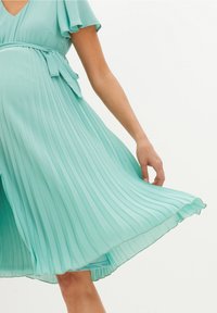 Mátově zelené skládací šaty s výstřihem do V, krátkými rozšířenými rukávy a páskem v oblasti pasu, s volnou sukní a jemným lemováním.