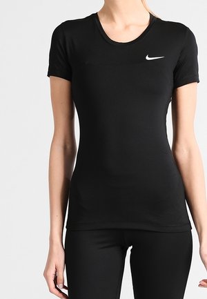 Frau trägt ein eng anliegendes schwarzes kurzärmeliges Nike T-Shirt und schwarze Hose, steht vor einem schlichten hellen Hintergrund.