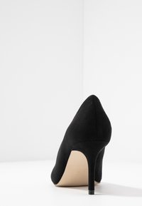 Escarpin à talons hauts en daim noir avec un bout pointu et un talon aiguille fin. Texture lisse et intérieur beige. Design simple et élégant.