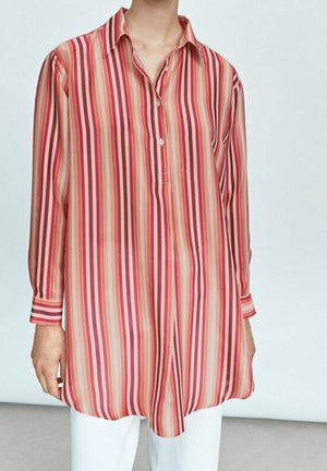 Personne portant une chemise à manches longues, coupe ample, avec des rayures verticales rouges, roses et beiges, associée à un pantalon blanc, sur un fond uni.