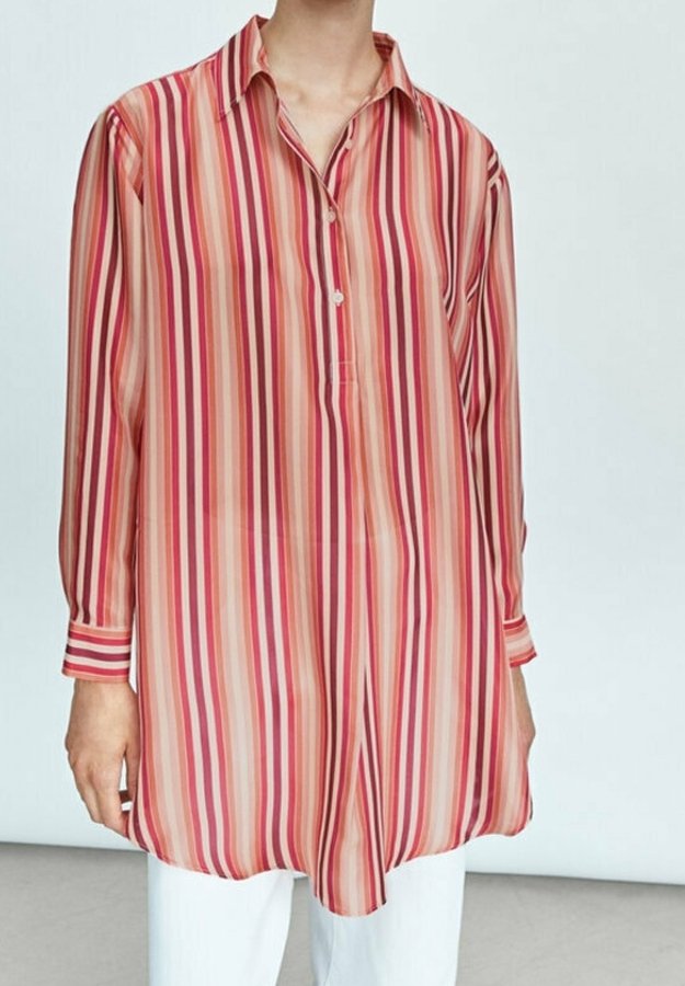 Personne portant une chemise à manches longues, coupe ample, avec des rayures verticales rouges, roses et beiges, associée à un pantalon blanc, sur un fond uni.