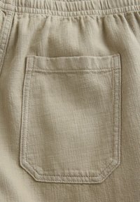 Beige fabric with a textured weave features an octagonal back pocket, reinforced stitching along the edges, and an elastic waistband.