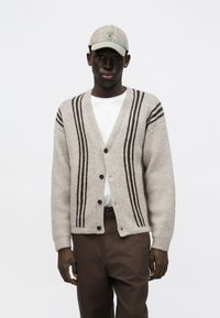 Szary kardigan z ciemnobrązowymi paskami, zapinany na guziki, o luźnym kroju. Noszony na białej koszuli, zestawiony z brązowymi spodniami i beżową czapką.