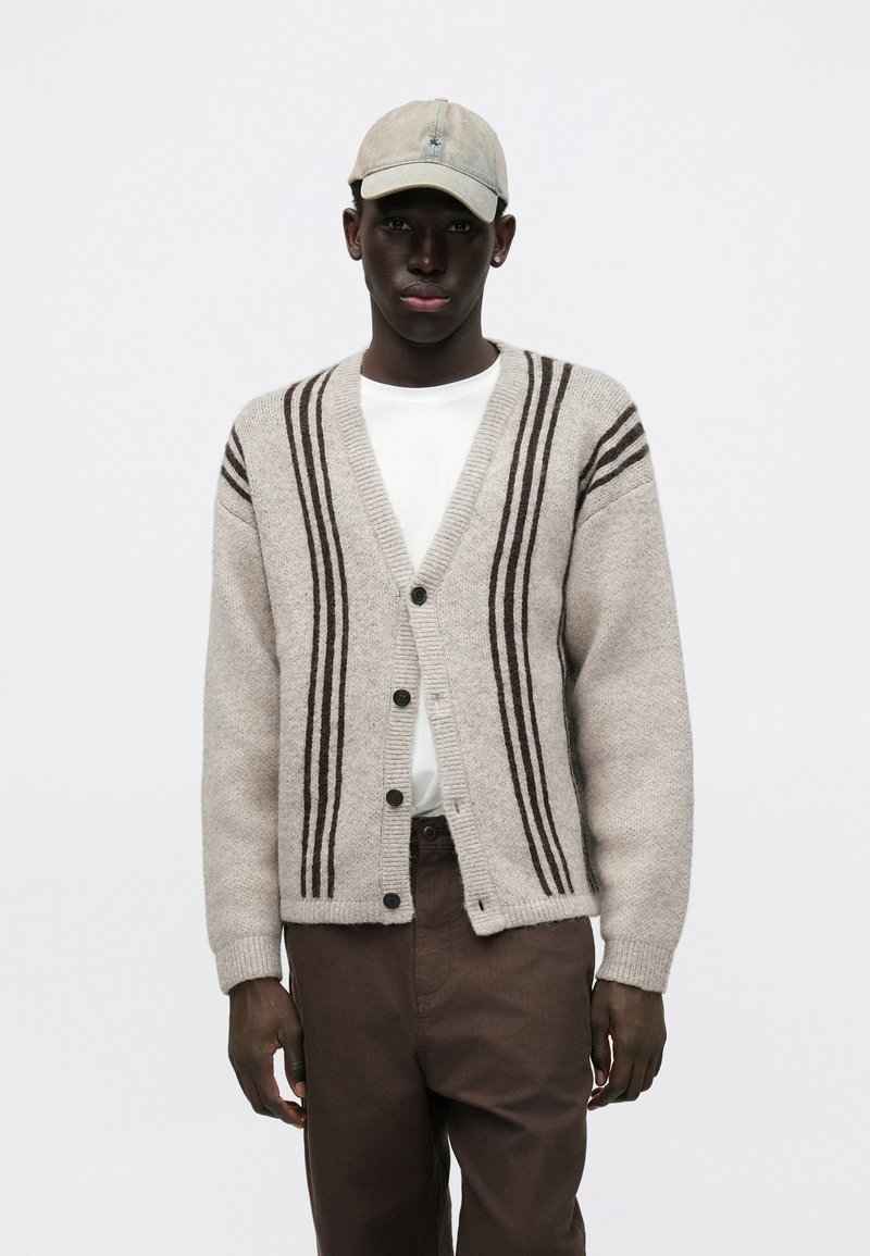 Szary kardigan z ciemnobrązowymi paskami, zapinany na guziki, o luźnym kroju. Noszony na białej koszuli, zestawiony z brązowymi spodniami i beżową czapką.