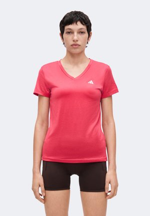 Kvinde med kort mørkt hår iført en lyserød V-hals T-shirt og sorte shorts, stående mod en ensfarvet lys baggrund.