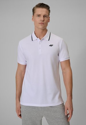 Mężczyzna ubrany w białą koszulkę polo z czarną lamówką i szare spodnie, stoi na jednolitym szarym tle, zwrócony przodem z neutralnym wyrazem twarzy.