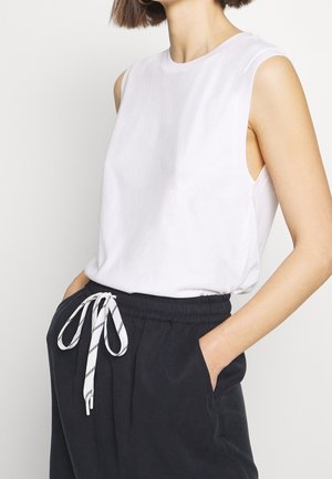 Top blanco sin mangas hecho de tela suave, con cuello redondo y un ajuste relajado, combinado con pantalones oscuros con un cordón de rayas.