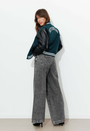 Femme portant une veste varsity verte avec des manches en cuir noir et le texte "SISTERSCLUB", associée à un jean large gris délavé à l'acide et des talons noirs.