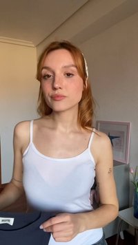 Giovane donna con capelli rossi e fascia bianca che indossa una canotta bianca, tiene un indumento blu scuro all'interno vicino a un quadro incorniciato e fiori.