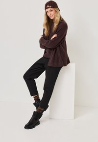 Tumepruun kudum kardigan, mustad püksid ja mustad pahkluudega saapad. Heledat pruunid leopardimustriga sokid. Model seisab valge pjedestaali vastu.