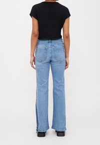 Persona che indossa jeans azzurri chiari con pannelli laterali scuri, una maglietta nera a maniche corte e scarpe nere con tacco, vista da dietro su uno sfondo bianco.