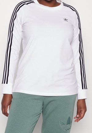 Personne portant un t-shirt Adidas à manches longues blanc avec des bandes noires sur les manches et un pantalon vert avec le logo Adidas sur la cuisse.