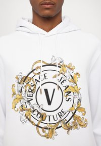 Balta hoodie su dideliu apvaliu juodu ir auksiniu grafiniu logotipu, papuoštu sudėtingais gėlių raštais ir tekstu "VERSACE JEANS COUTURE."