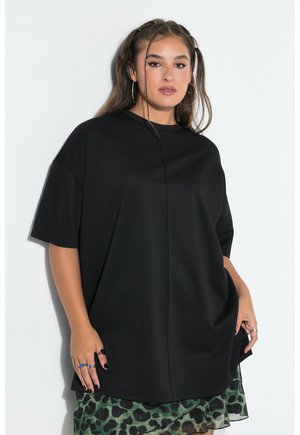 Kvinde iført oversized sort T-shirt over grøn og sort leopardmønstret nederdel, stående mod en hvid baggrund.