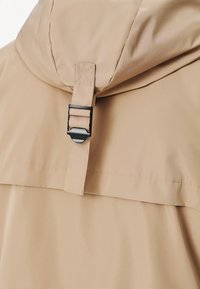 Jasno beżowa wodoodporna kurtka z kapturem, z dużym zapięciem na klamrę i gładką fakturą. Widoczne przeszycia dodają szczegółów.
