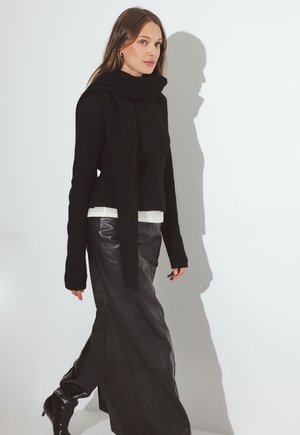 Mujer caminando con suéter negro abotonado, bufanda larga, falda de cuero negra y botas negras sobre fondo blanco.